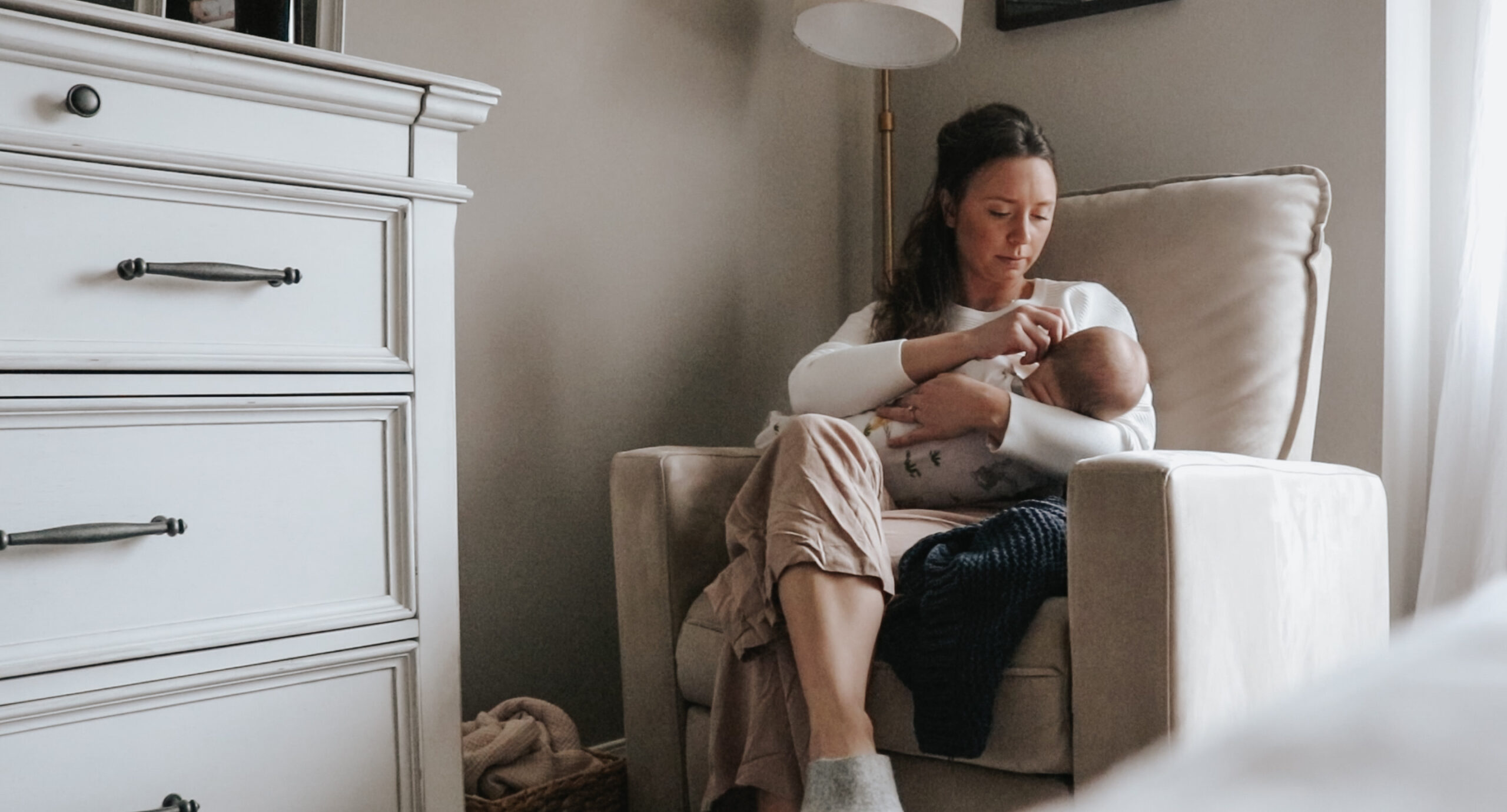 Homemaker and mom sit in a rocking chair while holding her baby. Her legs are crossed and she is touching the baby's face.